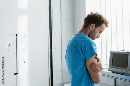 sad man in patient gown standing with crossed arms in hospital