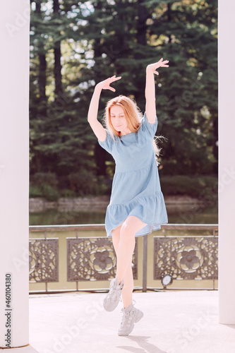 Fototapeta Naklejka Na Ścianę i Meble -  A long-haired lady in a short dress is posing in a summer park, standing on the toe of one leg and spreading her arms up. Ballet pose
