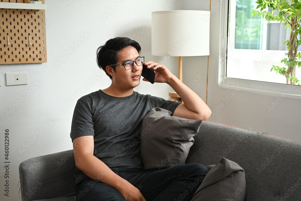 Photo of a young man on a sofa is talking on a mobile phone with a comfortable living room as a background. Photo of a young man on a sofa is talking on a mobile phone with a comfortable living room as a background.