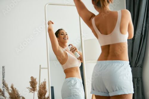 Young happy woman uses deodorant spray on her underarm while looking herself in a mirror.