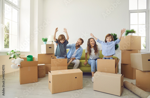 Happy young family having fun in their new home. Cheerful mommy and daddy sitting on couch and laughing while little kids are playing with cardboard boxes in spacious living room in new apartment