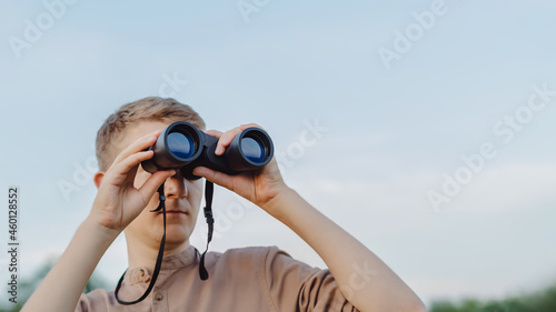 A man with modern binoculars against the sky and green forest.The concept of hunting, travel and outdoor recreation. Banner with copy space.A traveler or hunter is observing through binoculars.