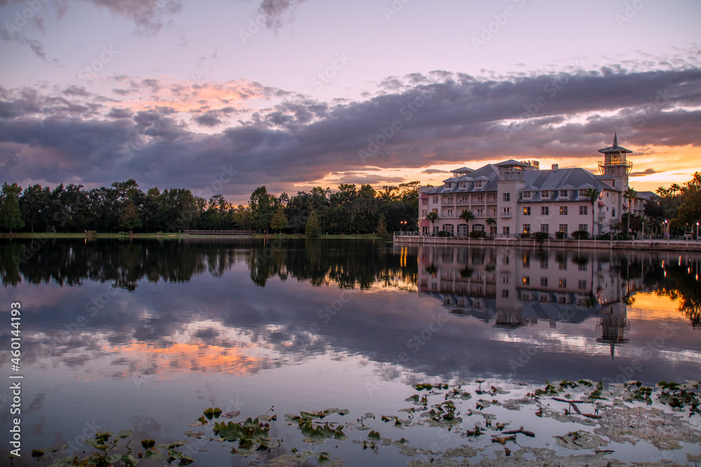 A hotel in Celebration (Orlando, Kissimmee), Florida reflects at the edge of a small lake during sunset with interesting cloud formations above.