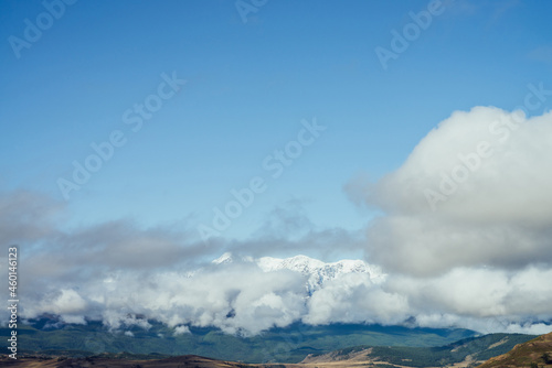 Awesome alpine landscape with great snowy mountain range in big low clouds under blue sky. Scenic view to snowy ridge above forest in large low cloud. Colorful mountain scenery with high mountains.