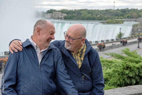 An elderly gay couple sit on a stone wall with Niagara Falls in the background.  They are looking at each other and laughing.  One has his arm around the shoulder of his husband.