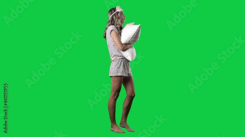 Side view of young woman in sleepwear with sleep mask holding pillow. Full body on green screen chroma key background