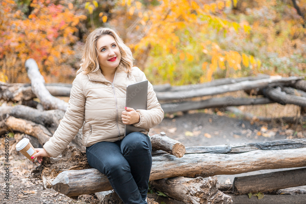 a beautiful girl is sitting on logs in an autumn park holding laptops and cups of coffee in her hand