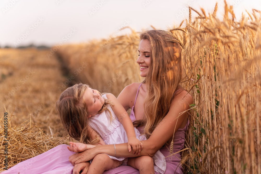 Mother and little daughter have fun sitting by the golden wheat field. Young woman tickles the girl Mother and little daughter have fun sitting by the golden wheat field. Young woman tickles the girl