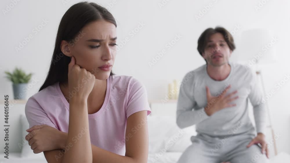 Relationship crisis. Close up portrait of young tired woman sitting on bed, excited man quarreling to her on background Relationship crisis. Close up portrait of young tired woman sitting on bed, excited man quarreling to her on background