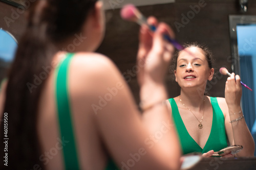 Mujer joven atractiva maquillándose la cara con artículos de belleza y maquillaje frente al espejo del cuarto de baño mientras sonríe para celebrar la nochebuena en familia