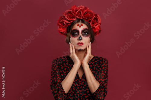 Beautiful girl in la muerta attire and skull make up looking to camera during halloween isolated on pink background.