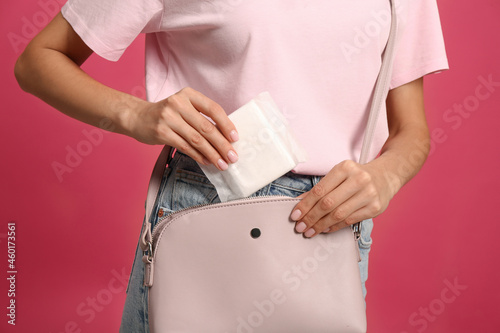 Young woman putting menstrual pad into purse on bright pink background, closeup