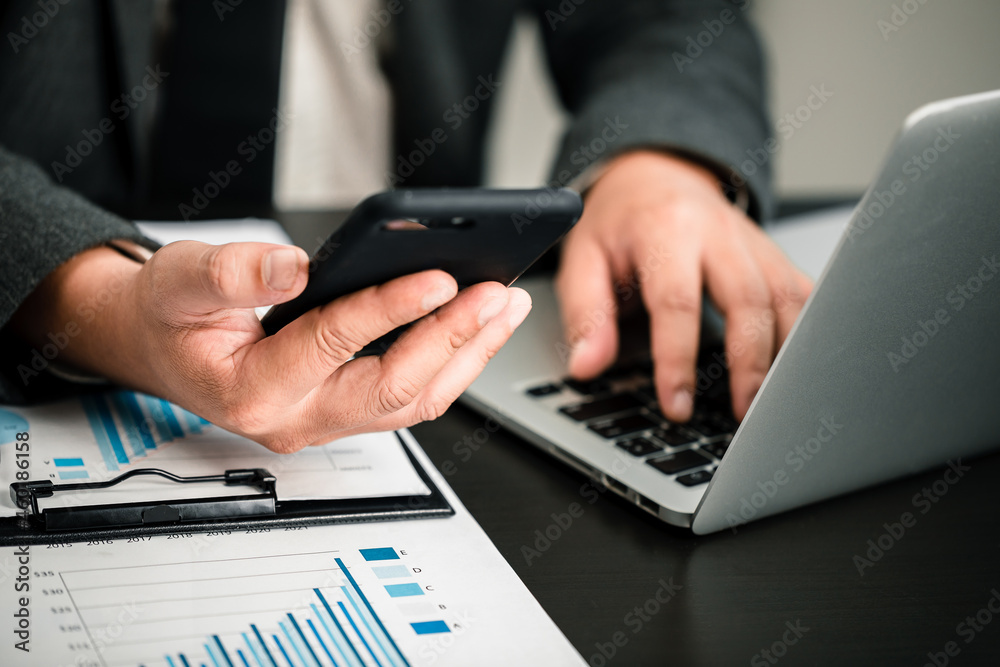 Fototapeta premium Close up of businessmen using smartphone and laptop computer working business report analytic at home, accounting document, marketing and financial business concept.