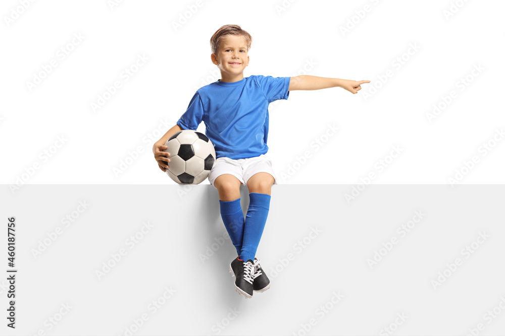 Boy in a blue soccer jersey sitting on a blank panel with a ball and pointing to the side