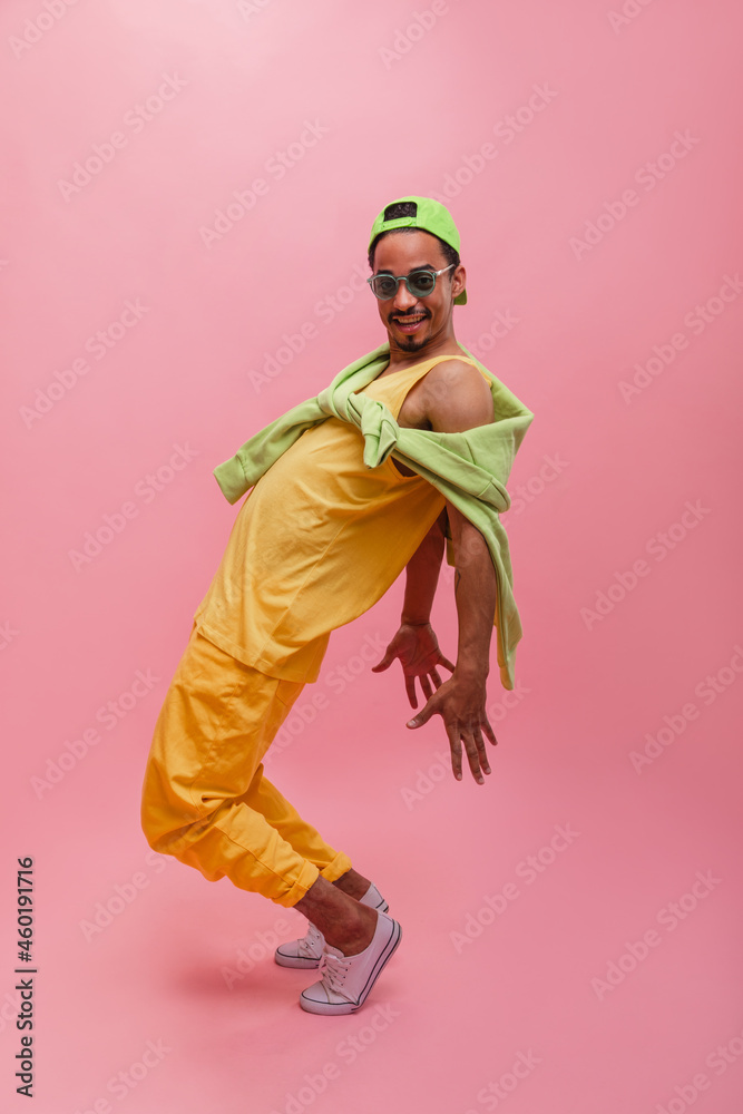 Full shot active african man standing on tiptoe, holding hands behind back with copy space. Attractive guy model with dark hair in summer yellow suit, glasses and cap. Lifestyle, male beauty concept Full shot active african man standing on tiptoe, holding hands behind back with copy space. Attractive guy model with dark hair in summer yellow suit, glasses and cap. Lifestyle, male beauty concept