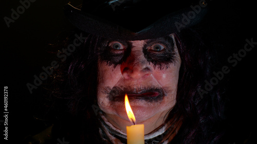 Sinister old elderly woman with horrible scary Halloween witch makeup in costume looking ominous at camera, making voodoo magic rituals with candle. Vampire granny isolated against black background