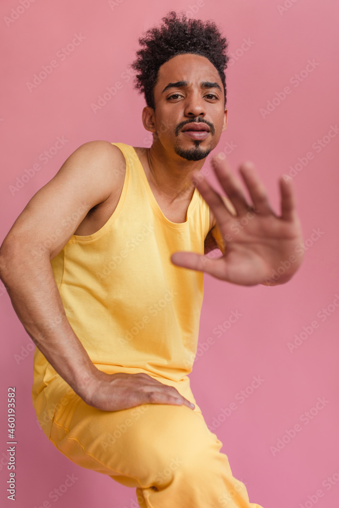Timid young swarthy American boy pushes camera away from him on pink background. Medium shot wearing light lemon tank top and yellow pants, looking straight into camera in studio. Timid young swarthy American boy pushes camera away from him on pink background. Medium shot wearing light lemon tank top and yellow pants, looking straight into camera in studio.