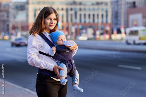 Mother is feeding baby in sling with bottle on street. © grigvovan