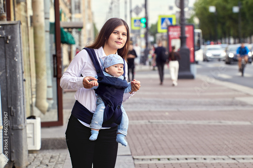 Young woman with baby in sling stands on crowded street in city.