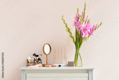 Vase with beautiful gladiolus flowers, female accessories and cosmetics on table against color wall