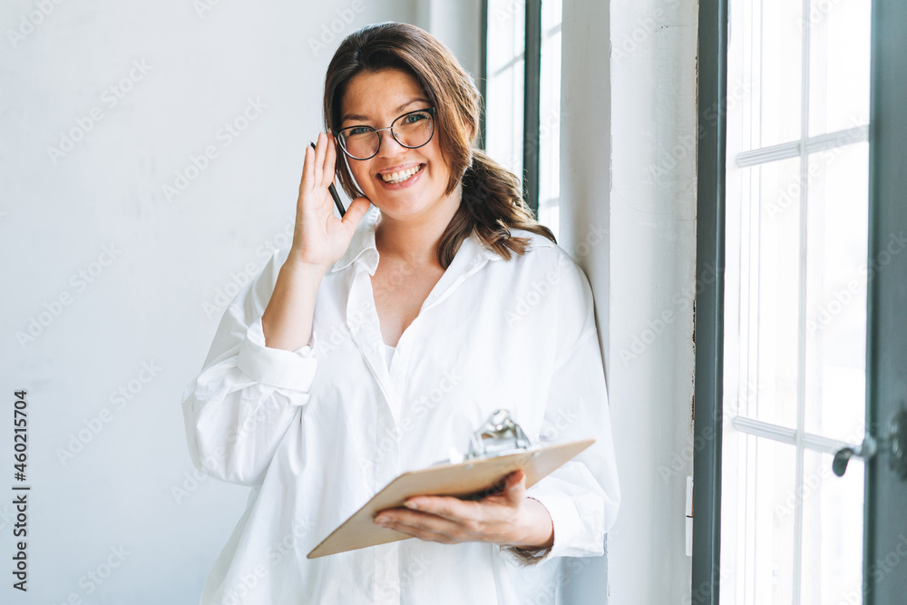 Young smiling brunette woman plus size in white shirt near window with pen and documents in hands in bright modern office Young smiling brunette woman plus size in white shirt near window with pen and documents in hands in bright modern office