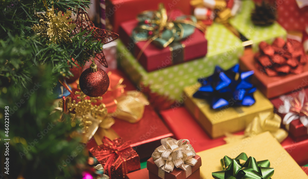 Close up shot of glossy red sphere ball hanging decorating on Christmas pine tree branch in front lot of Xmas eve wrapped present gift boxes stack with colorful ribbons and bows in blurred background