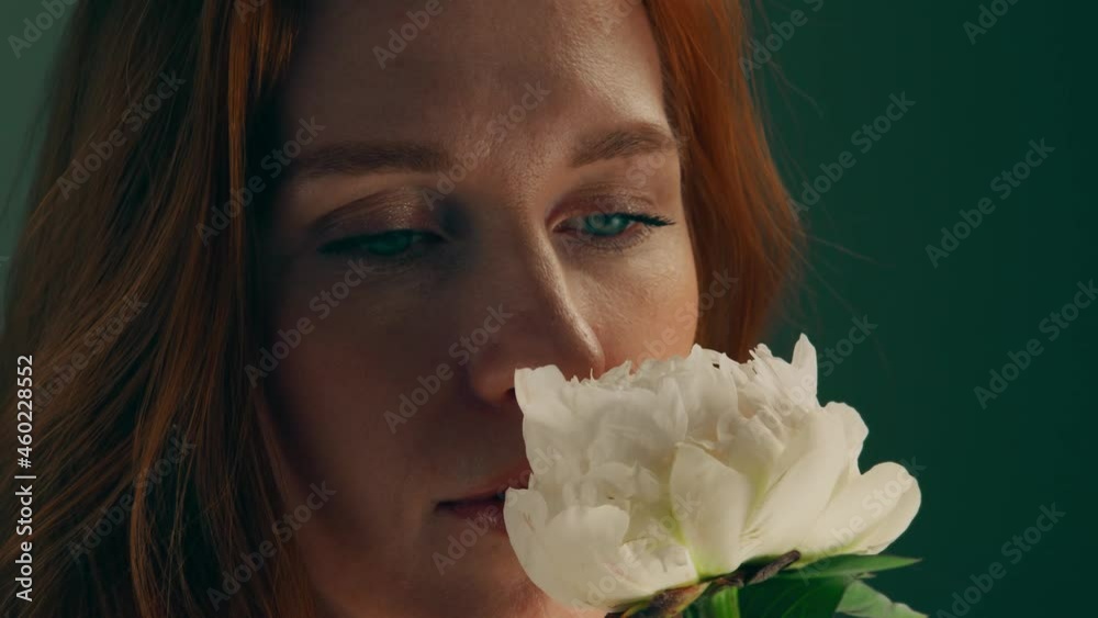 Close-up: A beautiful blue-eyed woman looks at a white flower. An attractive girl twirls a flower, enjoys the fragrance. The sun's glare reflects on the face. A charming red-haired woman. Slow motion Close-up: A beautiful blue-eyed woman looks at a white flower. An attractive girl twirls a flower, enjoys the fragrance. The sun's glare reflects on the face. A charming red-haired woman. Slow motion