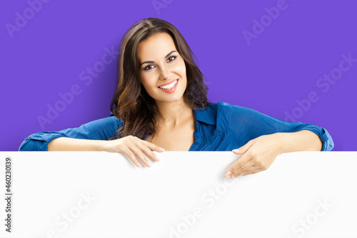 Happy smiling brunette young woman in blue clothing, standing behind, peeping from blank banner or showing mock up signboard with copy space for some text, over violet purple background.