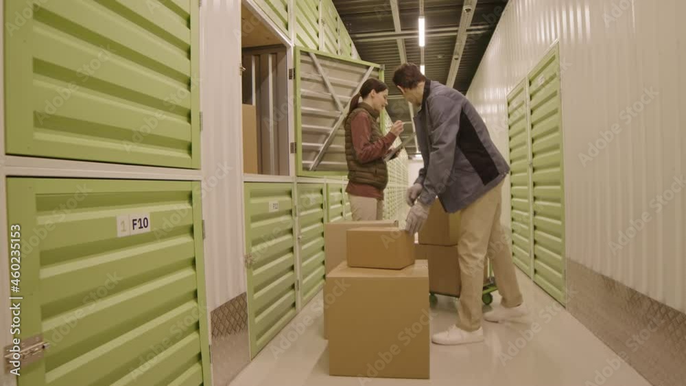 Wide shot of young woman and Caucasian man standing in hallway of warehouse, looking at cardboard boxes, keeping record of them
