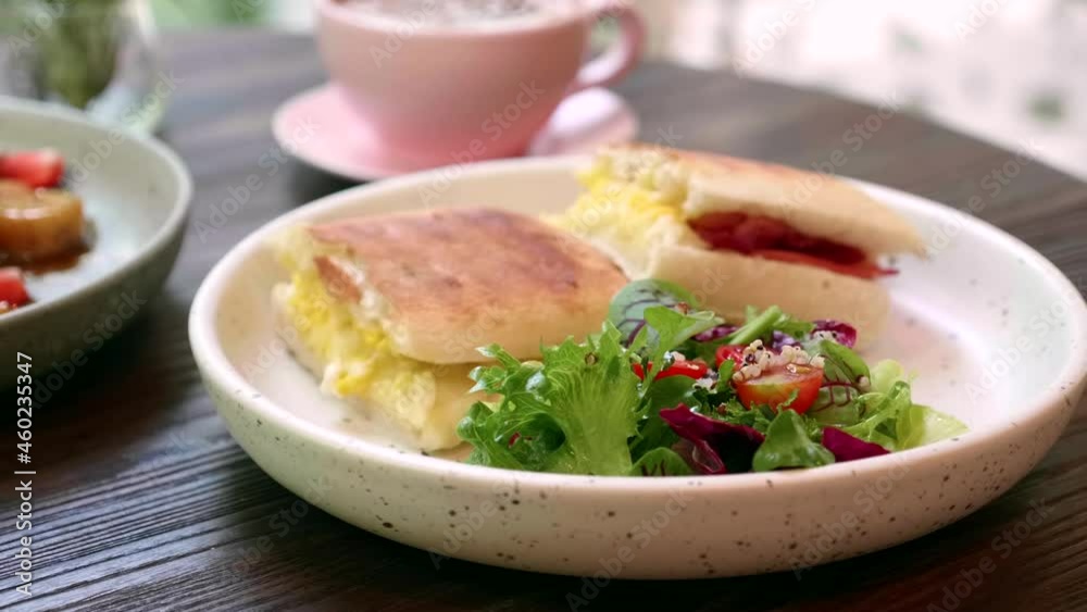 Sandwich with Egg, Cheese and Pepperoni with Fresh Green Salad on White Plate and Pink Cup of Coffee on Background. Breakfast or Lunch in Cafe. Delicious Fast Food on Wooden Table in Restaurant. Sandwich with Egg, Cheese and Pepperoni with Fresh Green Salad on White Plate and Pink Cup of Coffee on Background. Breakfast or Lunch in Cafe. Delicious Fast Food on Wooden Table in Restaurant.