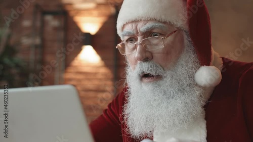 Close-up of white-bearded Santa Claus wearing eyeglasses, looking at cropped computer screen, getting shocked and surprised photo