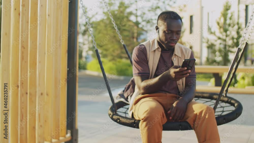 Young African American man sitting on round net swing in city and surfing the mobile internet on smartphone Young African American man sitting on round net swing in city and surfing the mobile internet on smartphone
