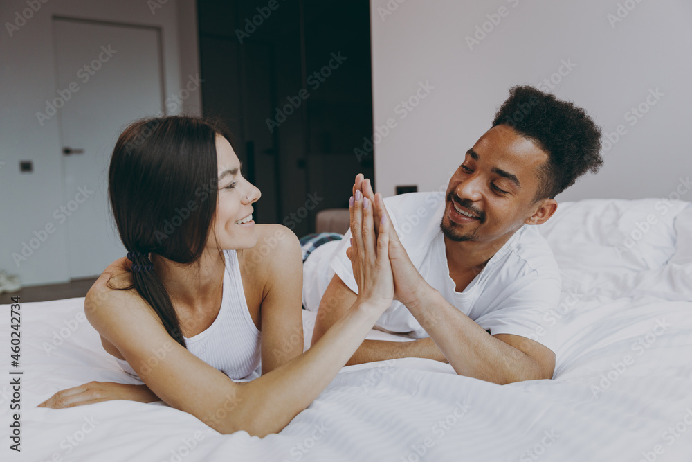 Fun young couple two man woman family in casual white clothes lying in bed on stomach give high five each other rest relax spend time together in bedroom lounge home house wake up be lost in reverie Fun young couple two man woman family in casual white clothes lying in bed on stomach give high five each other rest relax spend time together in bedroom lounge home house wake up be lost in reverie