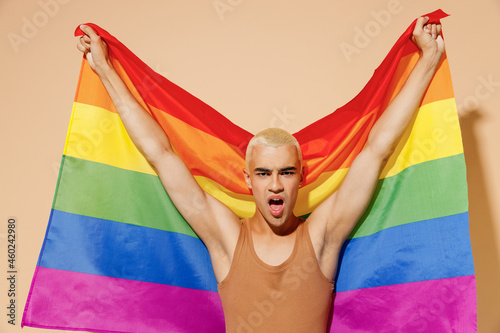 Young activist transsexual bisexual happy fun cool blond latin gay man with make up in beige tank shirt hold rainbow flag isolated on plain light ocher background studio People lgbt lifestyle concept.