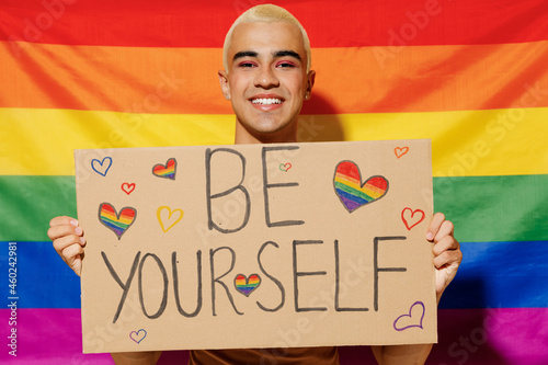 Young queer bisexual smiling blond latin gay man with make up in beige tank shirt hold card sign with be yourself title text on rainbow flag background studio portrait People lgbt lifestyle concept. photo