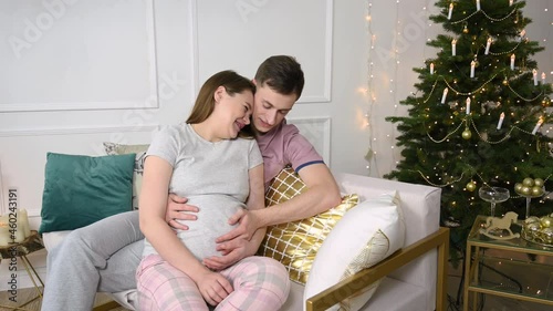 Caring husband talking to her pregnant wife's belly. Young pregnant couple resting on the sofa during Christmas time