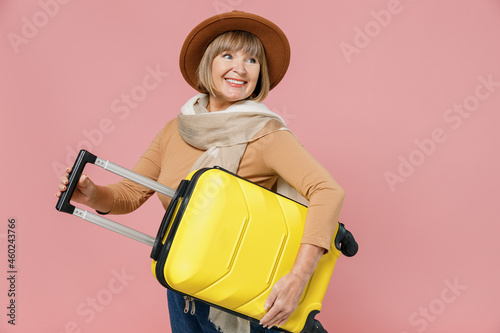 Traveler tourist mature elderly senior lady woman 55 years old wear brown shirt hat scarf hold under hand suitcase bag go move look back isolated on plain pastel light pink background studio portrait