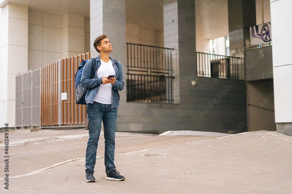 Full length portrait of handsome male courier with thermo backpack using navigation app on phone standing in city street. Delivery man searching for client address looking up. Full length portrait of handsome male courier with thermo backpack using navigation app on phone standing in city street. Delivery man searching for client address looking up.