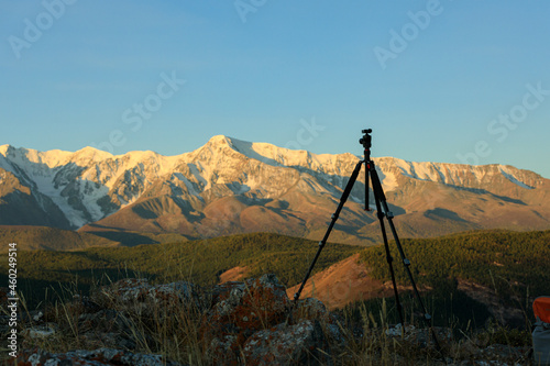 A tripod for a camera among the rocks against the background of high snowy peaks. Soft sunlight at sunset or dawn