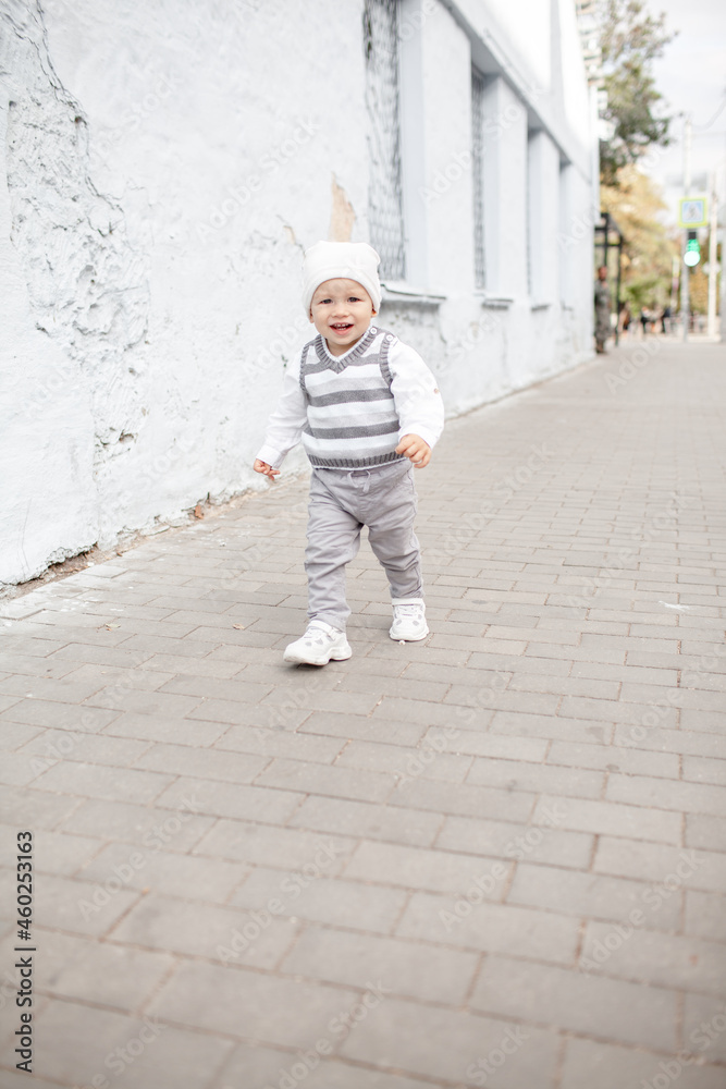 Stylish male toddler dressed fashion clothes, grey jeans, sweater and walking against the street wall
