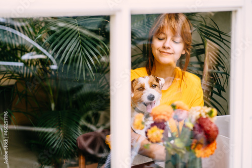 View through the window of caucasian girl student sitting at table with computer and dog at work place. Pretty amazing female freelancer working at cafe with jack russell terrier.