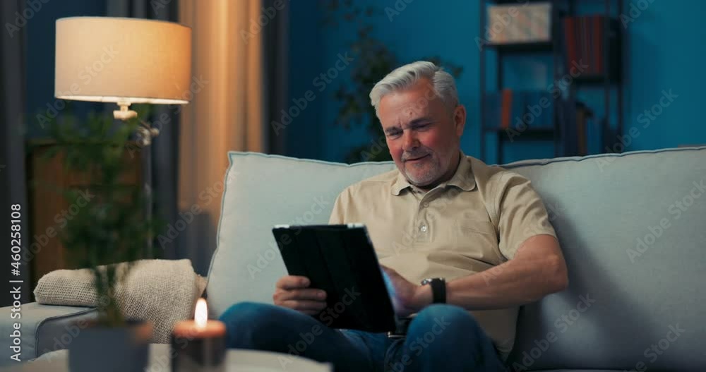 Smiling gray-haired man relaxing in living room on sofa with tablet. The retiree reads news on the internet, ebooks, weblogs. Guy learns how to use electronic devices, modern technology