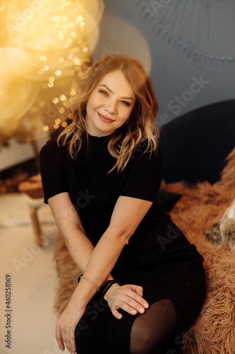 Portrait of stylish gorgeous woman sitting in cozy chair, smiling. Beautiful female in fashion clothes posing, looking directly at the camera, spending winter holidays at home, xmas concept
