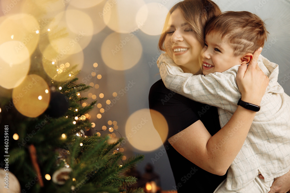 Overjoyed woman with little son hugging near Christmas tree, spending winter holidays at home. Beautiful mother celebrating xmas with adorable child, smile, enjoying happy moments, parenting concept Overjoyed woman with little son hugging near Christmas tree, spending winter holidays at home. Beautiful mother celebrating xmas with adorable child, smile, enjoying happy moments, parenting concept