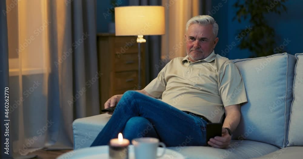 An elderly man with gray hair sits comfortably on sofa in evening room with leg tucked in, relaxing, resting, holding TV remote in hand, answering a message on phone in other hand, smiling An elderly man with gray hair sits comfortably on sofa in evening room with leg tucked in, relaxing, resting, holding TV remote in hand, answering a message on phone in other hand, smiling