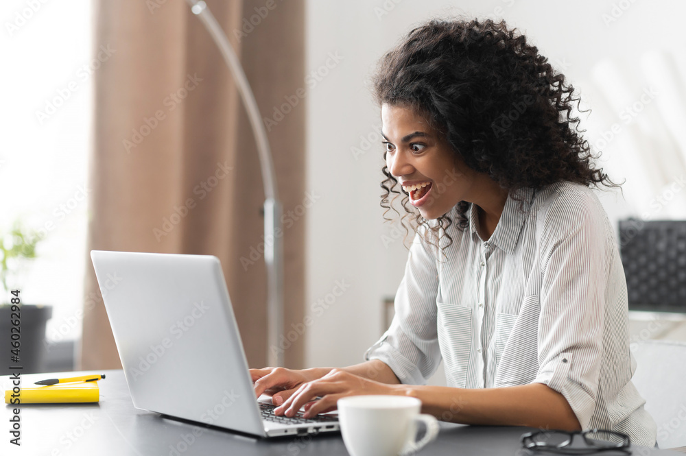Amazed African-American businesswoman in casual shirt sitting at the desk and staring at laptop screen, surprised mixed-race female office employee reading unexpected good news Amazed African-American businesswoman in casual shirt sitting at the desk and staring at laptop screen, surprised mixed-race female office employee reading unexpected good news