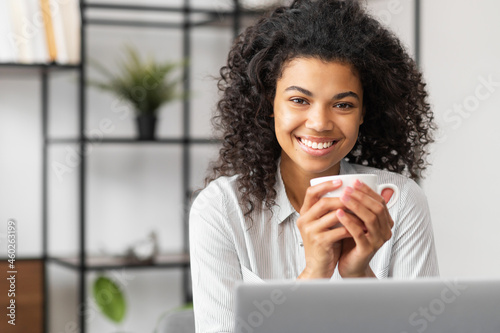 Cheerful happy African-American businesswoman in casual shirt sitting at the desk in front of laptop with mug in hands and looks at the camera, cheerful mixed-race woman enjoys morning coffee