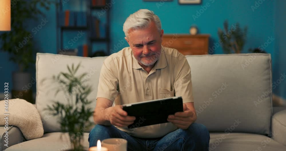 A man of mature age is relaxing in evening on sofa in living room, operating modern equipment, using a tablet, browsing news, reading emails, watching movies, happy fcet looks at camera with smile