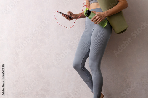 Woman wearing sportwear, holding bottle of water and gim mat,listening to music on smartphone against white empty wall.Free space