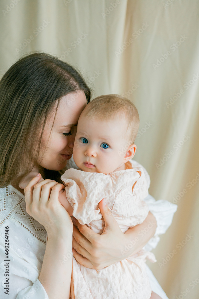 Closeup young beautiful mother in a white dress is holding her little blue-eyed daughter of 5 months in her arms. Motherhood.